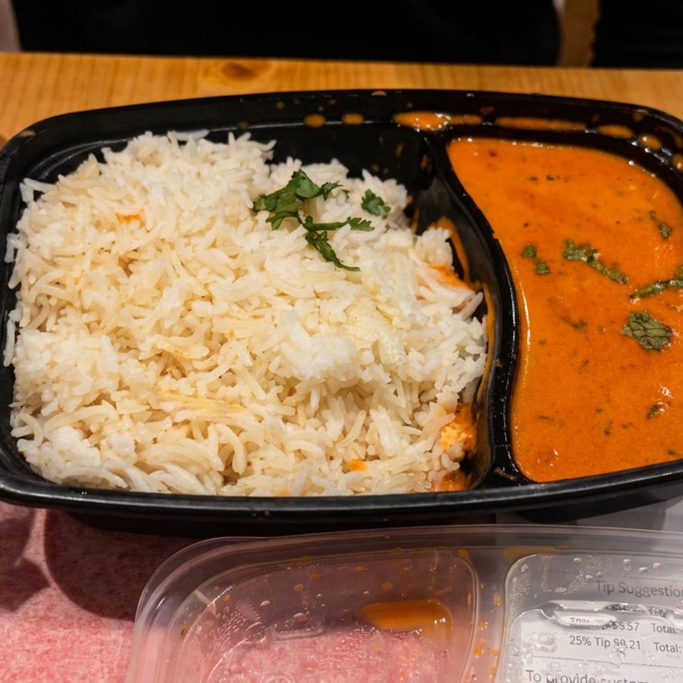 Black tray containing basmati rice garnished with cilantro and orange curry sauce on a wooden table, with foil-wrapped food and utensils nearby.